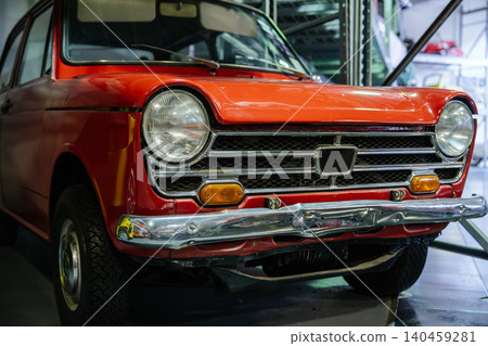 Detailed close-up captures front of vintage red car, emphasizing chrome grille, circular headlights, and dented bumper. Indoor setting features blurred industrial background elements. Detailed close-up captures front of vintage red car, emphasizing chrome grille, circular headlights, and dented bumper. Indoor setting features blurred industrial background elements. 140459281