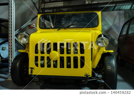 Close-up view captures a vintage yellow microcar parked indoors, showcasing its unique grille, round headlights, and vibrant color. Shelving units and another vintage car are visible in the background Close-up view captures a vintage yellow microcar parked indoors, showcasing its unique grille, round headlights, and vibrant color. Shelving units and another vintage car are visible in the background 140459282
