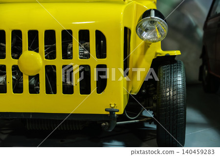 Detailed close-up captures front section of vibrant yellow vintage vehicle. Focus on distinctive grille and round headlight, with blurred background suggesting museum or showroom setting. Detailed close-up captures front section of vibrant yellow vintage vehicle. Focus on distinctive grille and round headlight, with blurred background suggesting museum or showroom setting. 140459283