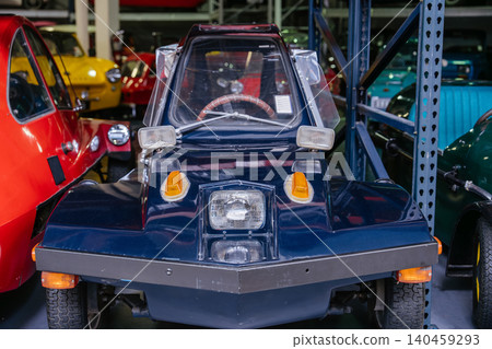 Close-up view of a distinctive dark blue three-wheeled microcar parked indoors, showcasing its bubble canopy and unique design. Vintage cars in red and yellow are partially visible in the background Close-up view of a distinctive dark blue three-wheeled microcar parked indoors, showcasing its bubble canopy and unique design. Vintage cars in red and yellow are partially visible in the background 140459293