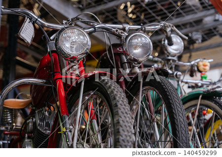 Close-up, low-angle view of vintage motorcycles lined up in workshop. Red motorcycle stands out with chrome handlebars and large headlight, while dark red and green motorcycles are partially visible. 140459299