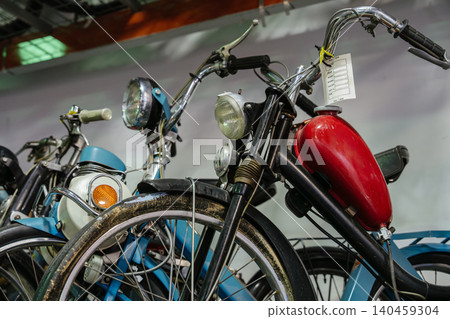 Close-up view captures several vintage mopeds lined up indoors. Prominent features include a bright red fuel tank and a blank information tag attached to handlebars, suggesting a display context. 140459304