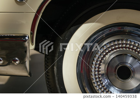Close-up captures intricate details of vintage car wheel featuring whitewall tire and diamond-patterned chrome hubcap. Chrome bumper showcases visible bolts, evoking a classic and luxurious feel. 140459338