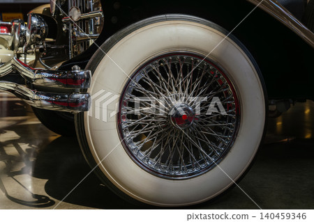 Detailed close-up captures vintage car's whitewall tire and chrome spoked wheel. Polished chrome hub and exhaust pipes reflect surroundings, showcasing classic automotive craftsmanship. 140459346