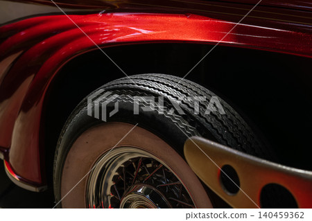 Detailed close-up showcases vintage car's wheel and fender. Vibrant metallic red paint highlights curves, while classic whitewall tire and chrome hub add elegance against a dark background. 140459362