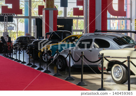 Vintage car exhibition showcases classic vehicles on a polished floor. A vibrant red carpet leads towards a lineup of antique cars, with palm trees visible through large windows. Vintage car exhibition showcases classic vehicles on a polished floor. A vibrant red carpet leads towards a lineup of antique cars, with palm trees visible through large windows. 140459365