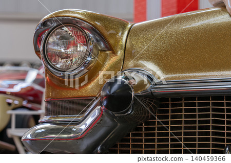 Detailed close-up captures front quarter of vintage automobile featuring shimmering gold glitter paint. Classic round headlight with chrome bezel and polished chrome grille create a striking design. 140459366