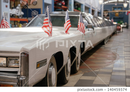 White stretch limousine parked indoors features three American flags on its hood. Tiled floor with checkered pattern and patriotic circular inlay enhances the luxurious atmosphere. 140459374