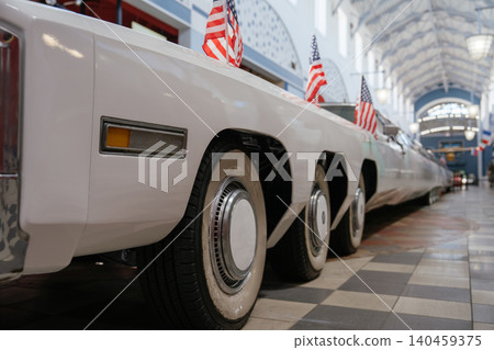Low-angle view captures side of long white limousine adorned with American flags. Vehicle is parked indoors in a building featuring checkered floor and arched ceiling, suggesting an exhibition setting Low-angle view captures side of long white limousine adorned with American flags. Vehicle is parked indoors in a building featuring checkered floor and arched ceiling, suggesting an exhibition setting 140459375