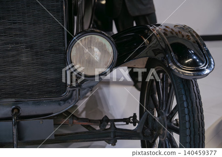 Detailed low-angle view highlights front section of vintage black automobile. Focus on classic design elements like horizontal radiator grille, round headlight, and curved fender over spoked wheel. Detailed low-angle view highlights front section of vintage black automobile. Focus on classic design elements like horizontal radiator grille, round headlight, and curved fender over spoked wheel. 140459377
