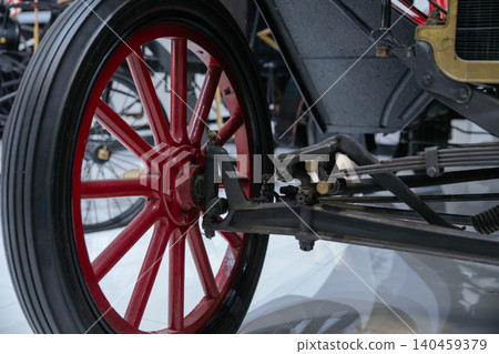 Detailed view showcases front wheel assembly of antique automobile. Bright red spoked rim and black tire highlight craftsmanship alongside dark metal and brass suspension components. 140459379