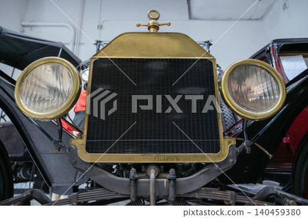 Close-up shot captures front view of vintage car, highlighting polished brass grille and large circular headlights. Mechanical components and museum setting add historical context. Close-up shot captures front view of vintage car, highlighting polished brass grille and large circular headlights. Mechanical components and museum setting add historical context. 140459380