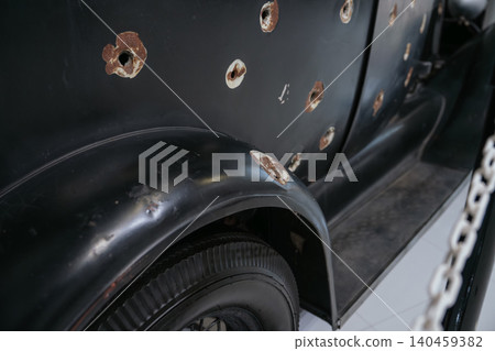 Close-up view of vintage black car body showing extensive bullet holes and rust. The damage highlights the car's history, with a chain-link fence suggesting a museum display setting. Close-up view of vintage black car body showing extensive bullet holes and rust. The damage highlights the car's history, with a chain-link fence suggesting a museum display setting. 140459382