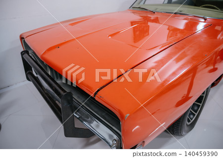 Detailed close-up view of vintage orange muscle car showcasing its front fender, hood, and grille with a black push bar. Minor scuffs on paint suggest a well-used vehicle in a garage setting. Detailed close-up view of vintage orange muscle car showcasing its front fender, hood, and grille with a black push bar. Minor scuffs on paint suggest a well-used vehicle in a garage setting. 140459390