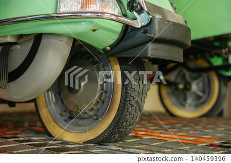 Detailed low-angle view captures front wheel and fender of vintage green scooter. Worn tire and tan sidewall are prominent, resting on metal grate with orange surface underneath. Detailed low-angle view captures front wheel and fender of vintage green scooter. Worn tire and tan sidewall are prominent, resting on metal grate with orange surface underneath. 140459396