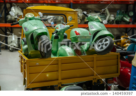 Several light green scooters are neatly packed in the cargo bed of a yellow flatbed truck. In the background, a variety of scooters in different colors are displayed on shelves, suggesting a storage Several light green scooters are neatly packed in the cargo bed of a yellow flatbed truck. In the background, a variety of scooters in different colors are displayed on shelves, suggesting a storage 140459397
