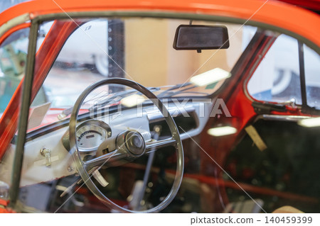 Detailed close-up view of vintage orange car interior, showcasing dashboard, steering wheel, and gauges. Reflections on windshield enhance nostalgic charm of classic automobile. Detailed close-up view of vintage orange car interior, showcasing dashboard, steering wheel, and gauges. Reflections on windshield enhance nostalgic charm of classic automobile. 140459399
