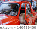 Close-up view of a bright red vintage microcar with an open driver's side door, showcasing checkered seat upholstery. Sunlight reflects off the polished exterior, emphasizing retro design in a 140459400