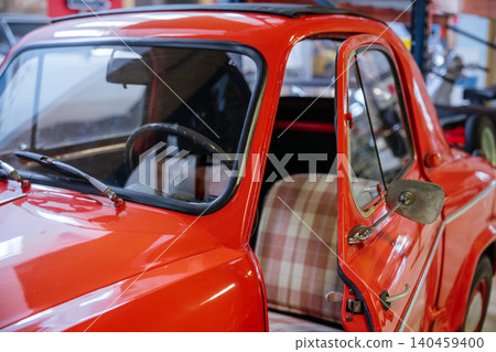 Close-up view of a bright red vintage microcar with an open driver's side door, showcasing checkered seat upholstery. Sunlight reflects off the polished exterior, emphasizing retro design in a Close-up view of a bright red vintage microcar with an open driver's side door, showcasing checkered seat upholstery. Sunlight reflects off the polished exterior, emphasizing retro design in a 140459400