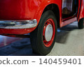 Detailed low-angle view captures rear wheel and bumper of classic red automobile. Shiny red paint contrasts with worn chrome bumper, showcasing retro automotive design aesthetics. 140459401