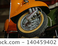 Detailed close-up captures front wheel and chrome suspension of vintage orange scooter. Soft lighting enhances textures, with a green scooter blurred in the background, suggesting a collection. 140459402