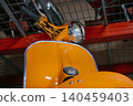 Close-up view captures vibrant orange vintage scooter against industrial shelving. Low-angle perspective emphasizes scooter's classic design and polished bodywork, reflecting workshop elements. 140459403