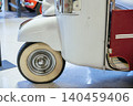 Detailed view of front section of vintage white three-wheeled vehicle, showcasing classic design elements. Prominent whitewall tire and shiny chrome detail on fender enhance retro appeal. 140459406