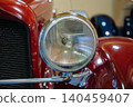 Detailed close-up view of classic red car's headlight and grill. Round headlight features ribbed glass lens and chrome trim, while polished red paint reflects surrounding light. 140459407