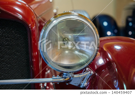 Detailed close-up view of classic red car's headlight and grill. Round headlight features ribbed glass lens and chrome trim, while polished red paint reflects surrounding light. Detailed close-up view of classic red car's headlight and grill. Round headlight features ribbed glass lens and chrome trim, while polished red paint reflects surrounding light. 140459407