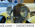 Detailed close-up view of front of vintage yellow car. Polished chrome grille and large round headlights with ribbed glass create a striking automotive design in an exhibition setting. 140459408