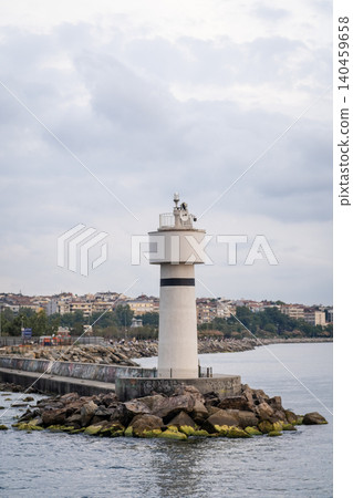 Bosphorus Lighthouse rises prominently from rocky coastline, surrounded by calm waters and distant city buildings under an overcast sky. Bosphorus Lighthouse rises prominently from rocky coastline, surrounded by calm waters and distant city buildings under an overcast sky. 140459658