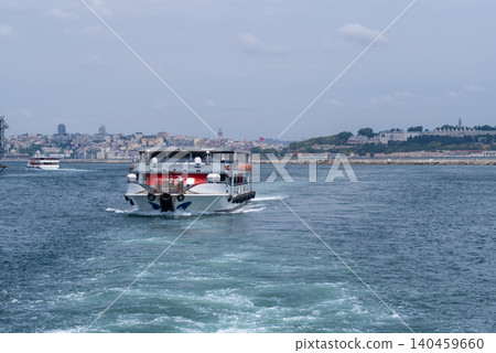 A ferry glides along the Bosphorus, creating ripples in the clear water, while the lively city landscape unfolds behind it under a bright sky 140459660