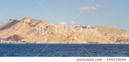Majestic golden mountains rise above a tranquil sea, with a small coastal settlement nestled at the foot, all under a serene afternoon sky. 140459694