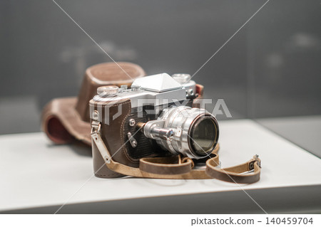 A vintage camera sits on display in a museum, showcasing its unique design with a leather strap and case, celebrating photographys past. 140459704