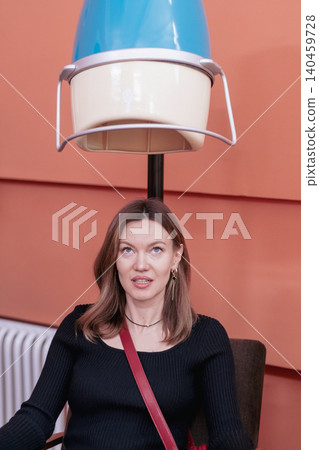 A woman with long hair relaxes in a salon chair under a blue and beige retro hair dryer, soaking up the ambiance of the bright interior environment. A woman with long hair relaxes in a salon chair under a blue and beige retro hair dryer, soaking up the ambiance of the bright interior environment. 140459728