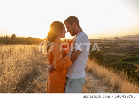 A couple enjoying a romantic moment as they embrace against a backdrop of orange sunset and rolling hills, surrounded by natures beauty. 140459763