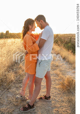 A couple enjoys a warm embrace as the sun sets, casting a golden hue over a tranquil outdoor path surrounded by dry grass and a scenic backdrop. 140459764