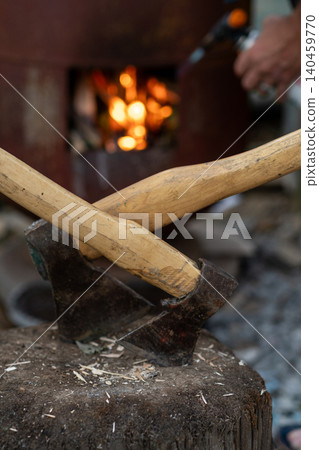 Two wooden handles cross over a black axe blade resting on a wooden stump, surrounded by shavings and a warm campfire glow in the background. 140459770