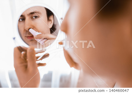 Man looking in a mirror using an electric trimmer for nose hair removal. A daily male grooming routine and personal facial care for health and hygiene. Man looking in a mirror using an electric trimmer for nose hair removal. A daily male grooming routine and personal facial care for health and hygiene. 140460047