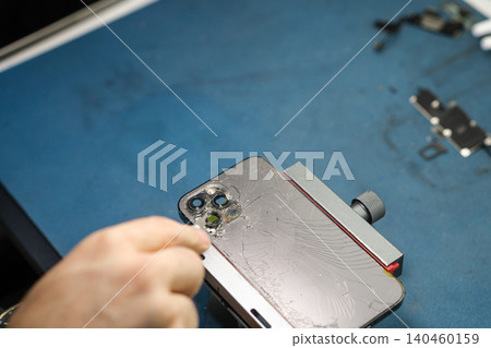 Person removes parts from a smartphone in a workshop. The process involves careful disassembly using tools on a blue surface. Person removes parts from a smartphone in a workshop. The process involves careful disassembly using tools on a blue surface. 140460159