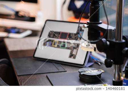 A magnifying device is set up on a workbench next to a tablet displaying circuit board designs. Tools are arranged nearby for electronics work. A magnifying device is set up on a workbench next to a tablet displaying circuit board designs. Tools are arranged nearby for electronics work. 140460181