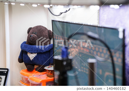 A brown teddy bear made from yarn stands on a desk beside a computer monitor. Various tools are placed nearby in a workspace during the day. A brown teddy bear made from yarn stands on a desk beside a computer monitor. Various tools are placed nearby in a workspace during the day. 140460182