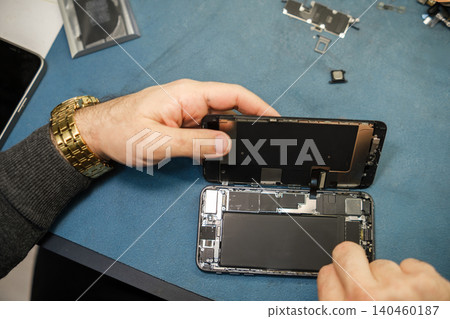 A technician works on a smartphone, carefully removing the back cover while tools and components are organized on a blue surface. A technician works on a smartphone, carefully removing the back cover while tools and components are organized on a blue surface. 140460187