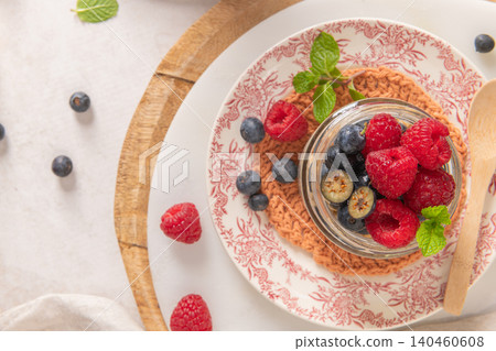 A close overhead shot shows a glass jar of overnight oats with chia seeds topped with fresh raspberries and blueberries on a floral plate with a wooden spoon and rustic kitchen elements 140460608