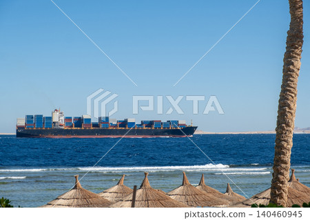 A large container ship glides over the blue sea, with palm trees and umbrellas on the sandy beach, creating a peaceful coastal scene. A large container ship glides over the blue sea, with palm trees and umbrellas on the sandy beach, creating a peaceful coastal scene. 140460945
