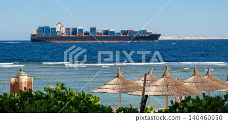 Container ship navigates through blue waters near a sandy beach with straw umbrellas under a bright sky, creating a picturesque coastal view. Container ship navigates through blue waters near a sandy beach with straw umbrellas under a bright sky, creating a picturesque coastal view. 140460950