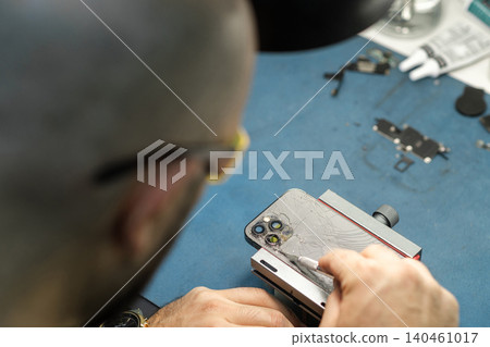 A technician is focused on fixing a smartphone screen in a repair workshop during the afternoon. Tools and parts are visible on the table. A technician is focused on fixing a smartphone screen in a repair workshop during the afternoon. Tools and parts are visible on the table. 140461017