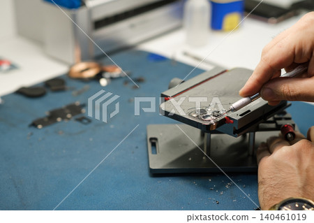 A technician uses a tool to repair an electronic device on a workbench cluttered with parts and tools in a workshop setting. A technician uses a tool to repair an electronic device on a workbench cluttered with parts and tools in a workshop setting. 140461019