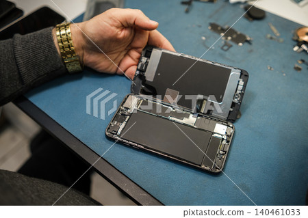 A person is repairing a smartphone by holding the device with the screen detached on a workbench. Tools and parts are visible. 140461033