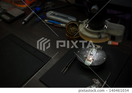 A person inspects a circuit board using a magnifying glass while surrounded by tools in a workspace during daytime. A person inspects a circuit board using a magnifying glass while surrounded by tools in a workspace during daytime. 140461034